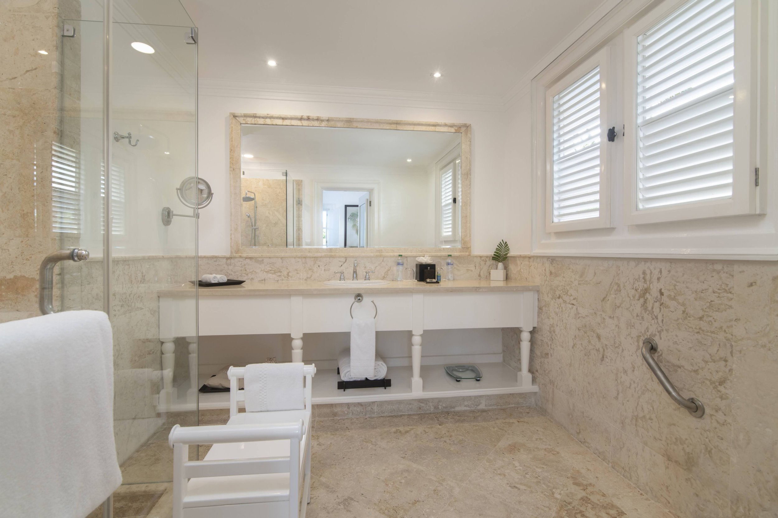 Bright bathroom with large mirror, beige marble walls and floor, white vanity, and two windows with white shutters.