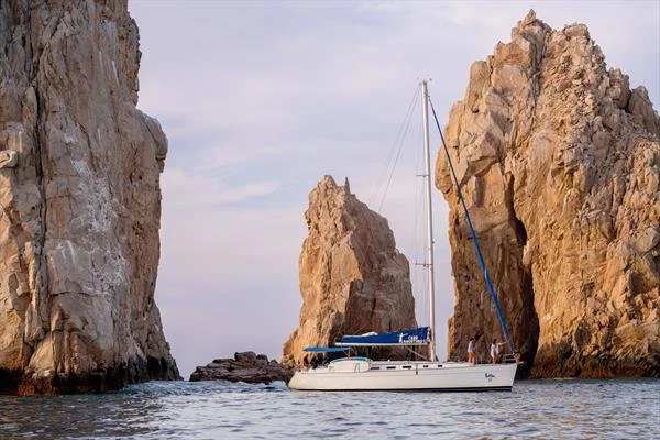 Sailboat navigating through a narrow passage between large rocky cliffs with a cloudy sky above.