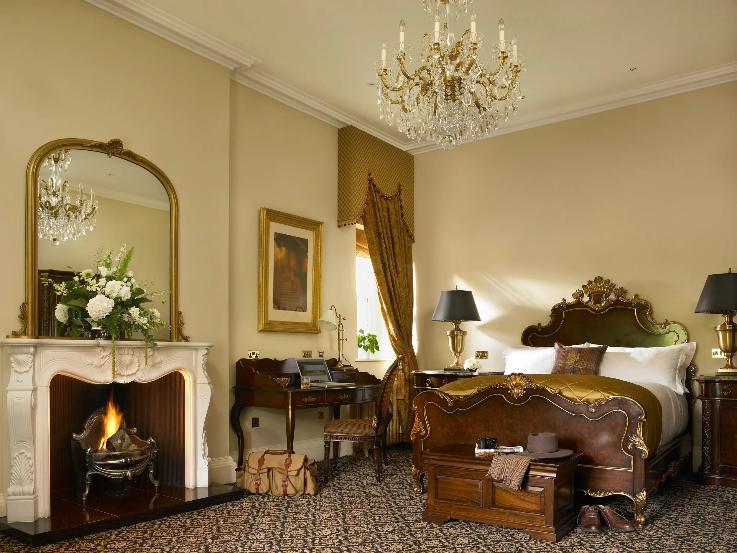 A luxurious hotel bedroom with a bed, bedside tables, and chandeliers. The fireplace has a bouquet of white flowers, and there are various personal items including a camera, hat, and shoes on the furniture.