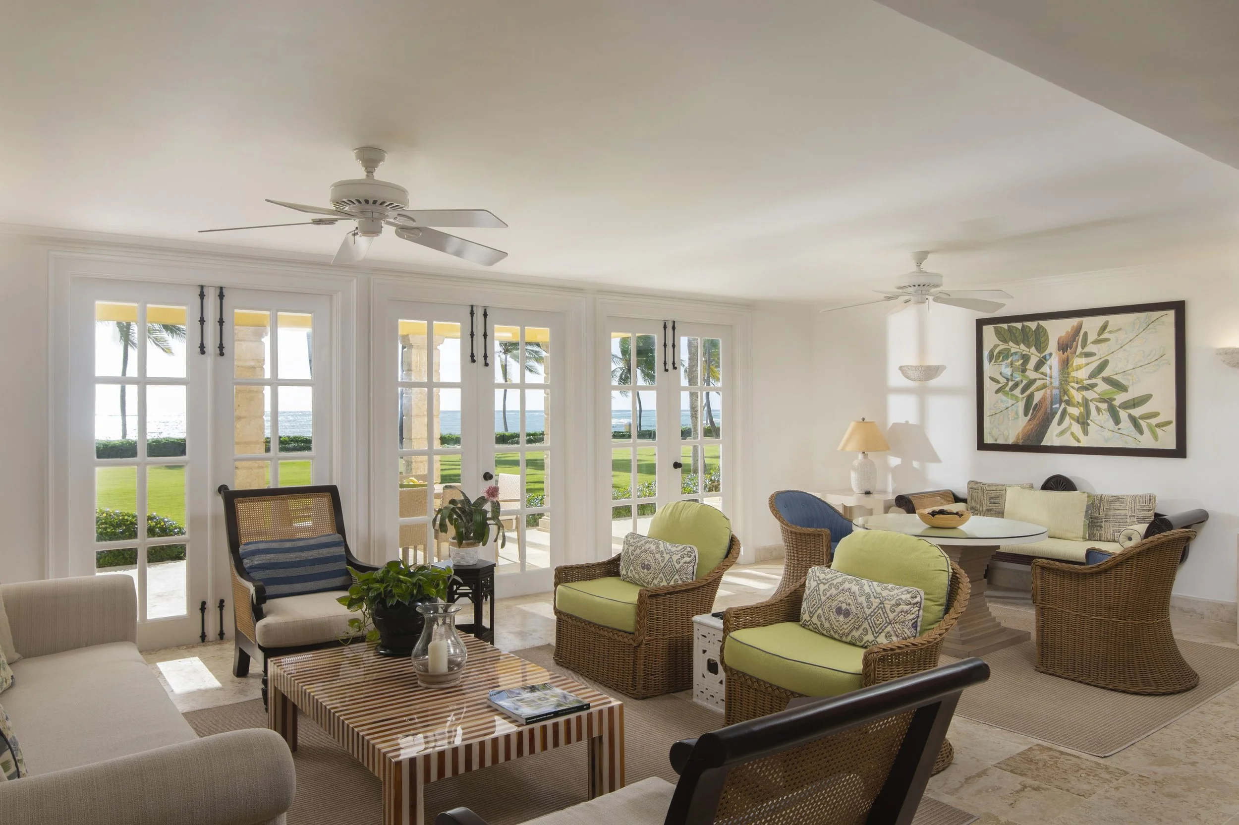Living room with large glass doors, wicker and upholstered chairs, a round table, potted plants, wall art, ceiling fans, and a view of palm trees and the ocean outside.