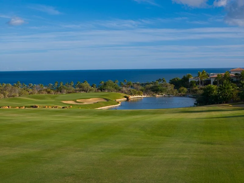 A golf course with green fairways, sand bunkers, and a water hazard, overlooking the ocean under a partly cloudy sky.