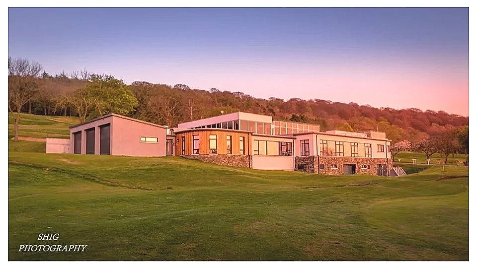 A modern residential house with large windows, situated on a grassy hill with a backdrop of trees and a colorful sky at sunset.