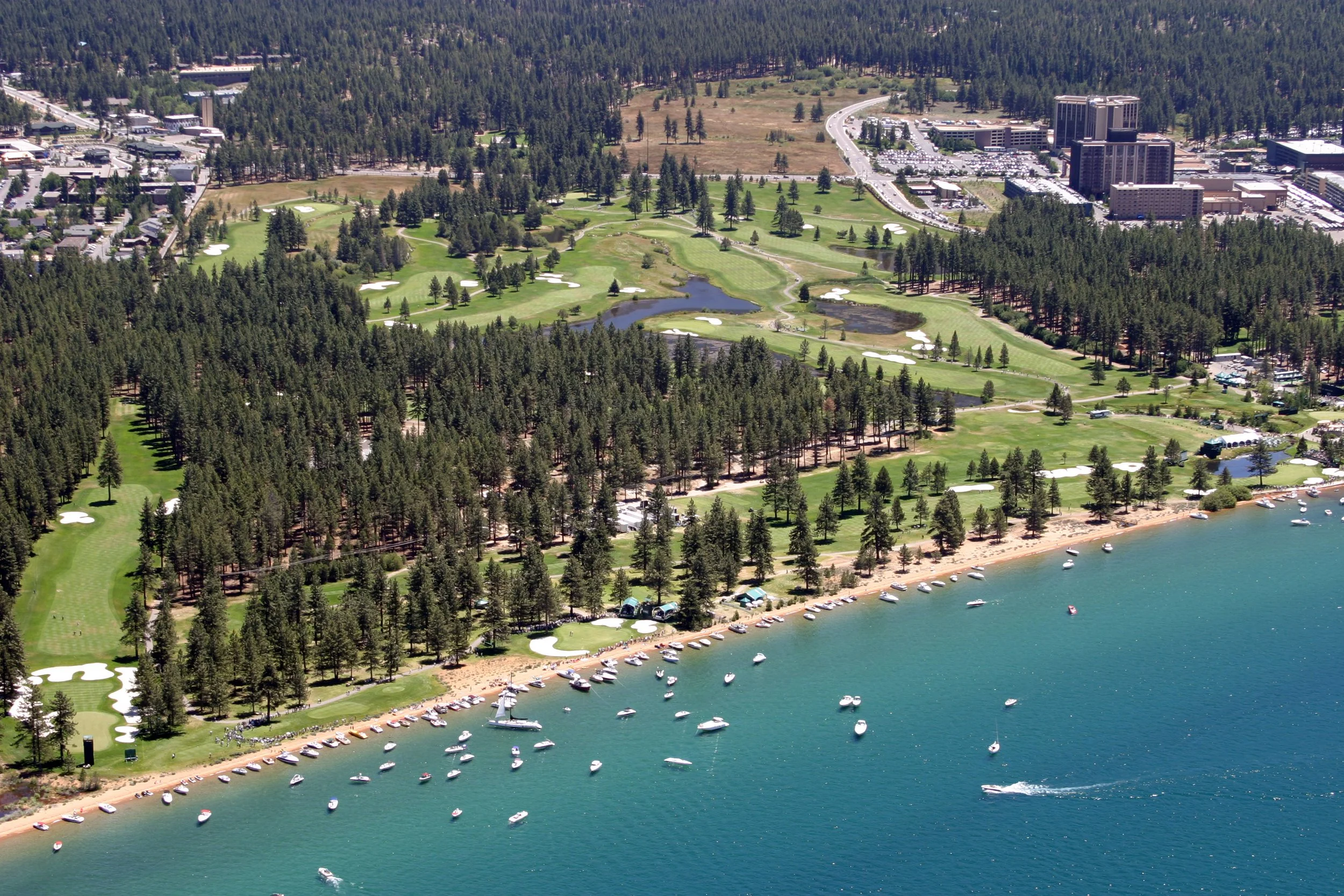 Aerial view of a golf course adjacent to a lake with boats, surrounded by dense forest and city buildings in the background.