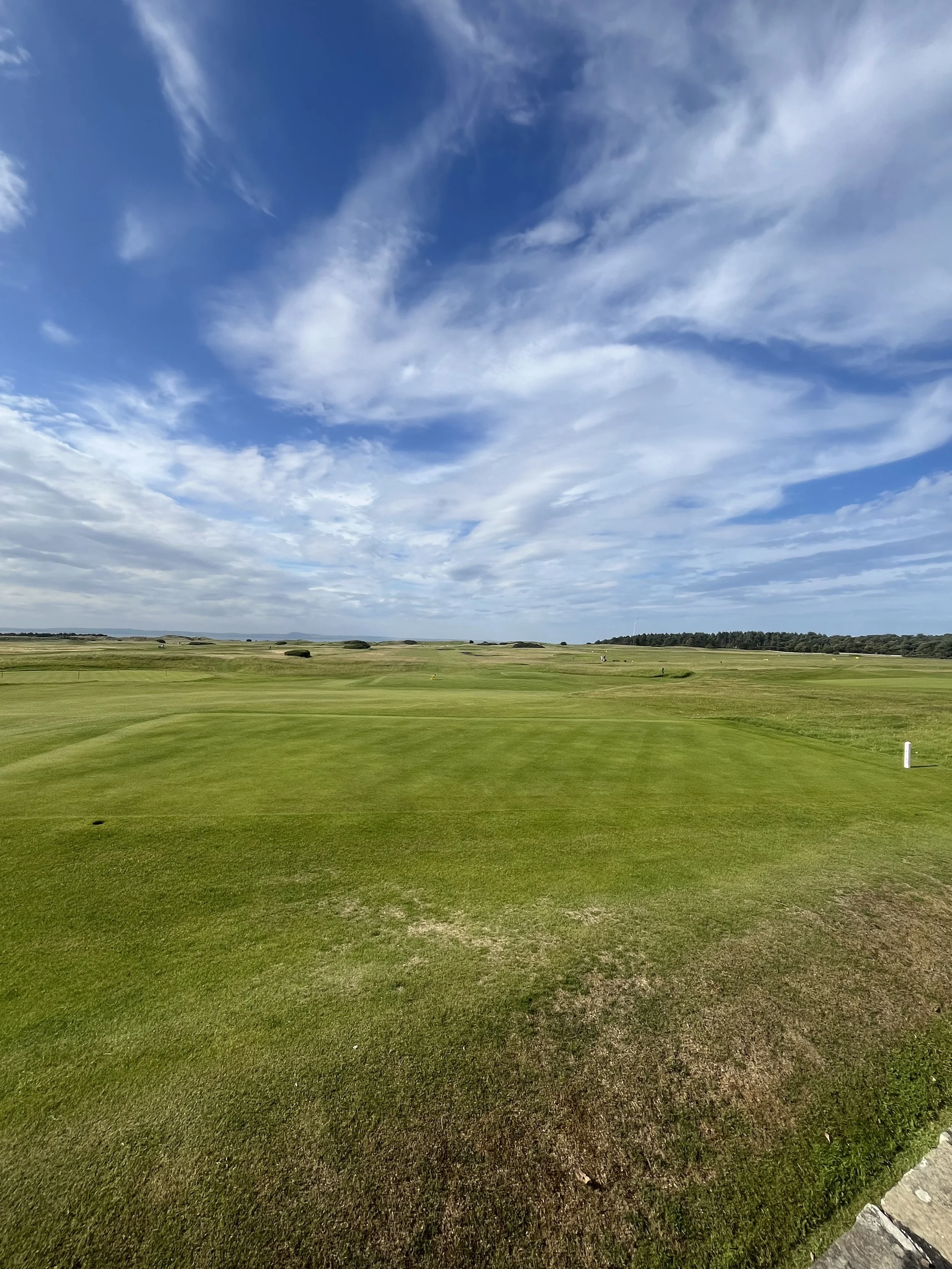 A wide view of a golf course with green grass, fairways, and a mostly blue sky with scattered clouds.
