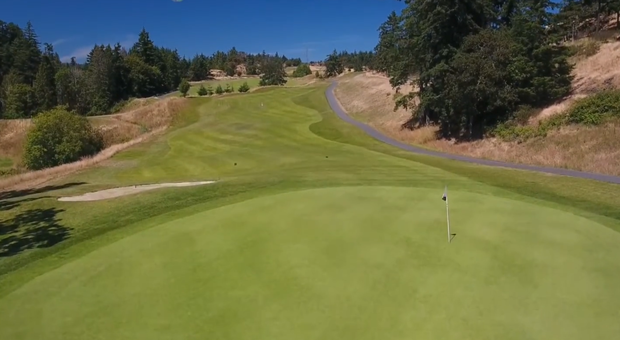 A golf course green with a flagstick and hole, surrounded by well-maintained grass and trees, with a paved path running along the right side and a rolling hillside in the background.