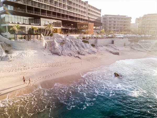 Two people walking along the sandy beach at sunset, with modern buildings and palm trees in the background. Waves are gently crashing onto the shore.