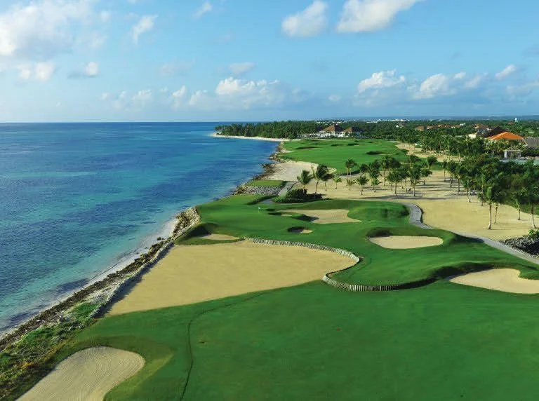Aerial view of a coastal golf course with green fairways, sand bunkers, and a rocky shoreline along the ocean under a partly cloudy sky.
