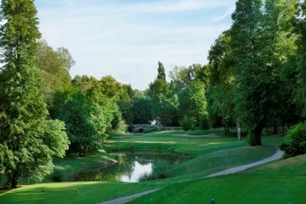 A scenic golf course with a water hazard, surrounded by lush green trees and a clear blue sky.