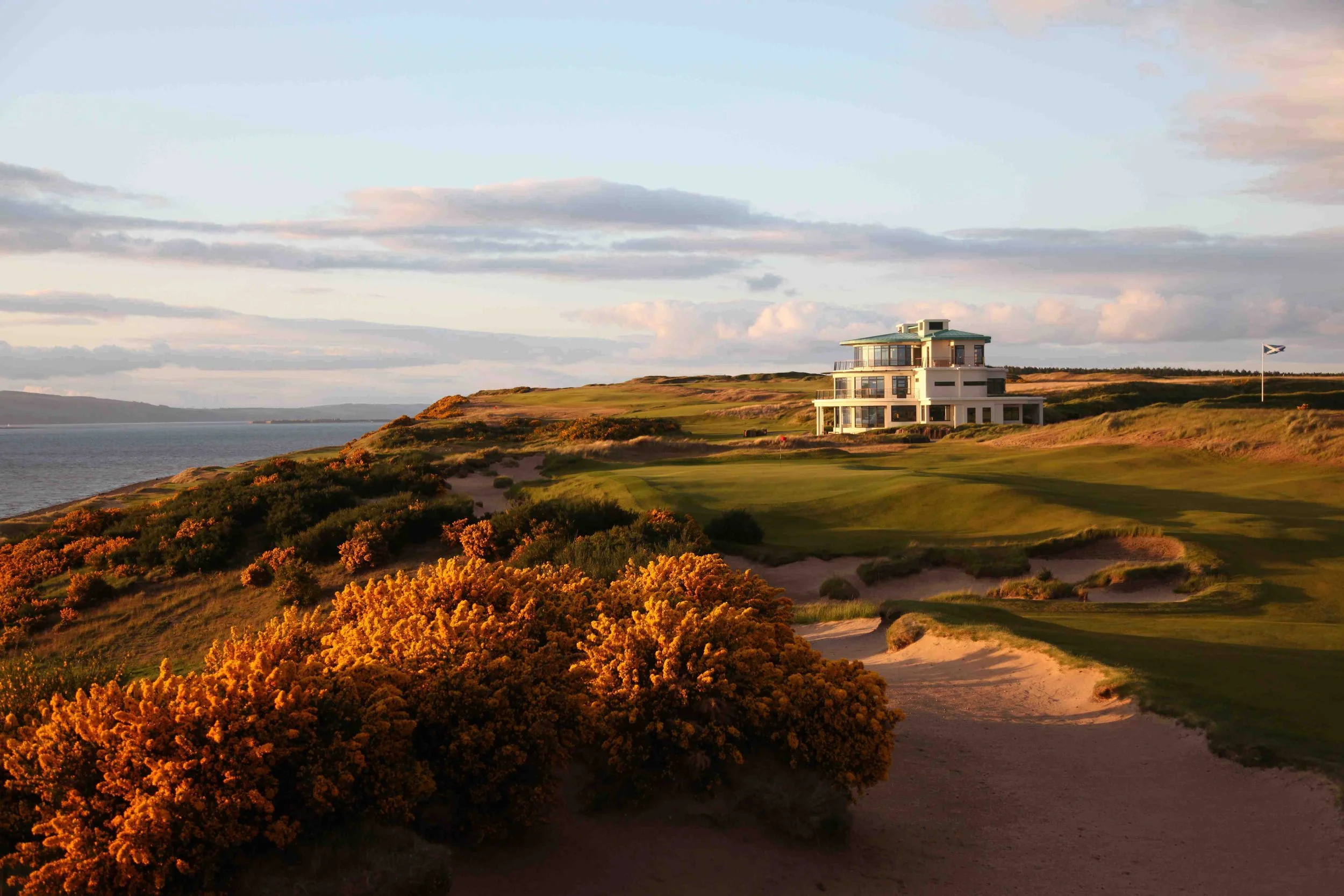 A seaside golf course with a modern house on a hill, surrounded by bushes and sand dunes, under a partly cloudy sky.