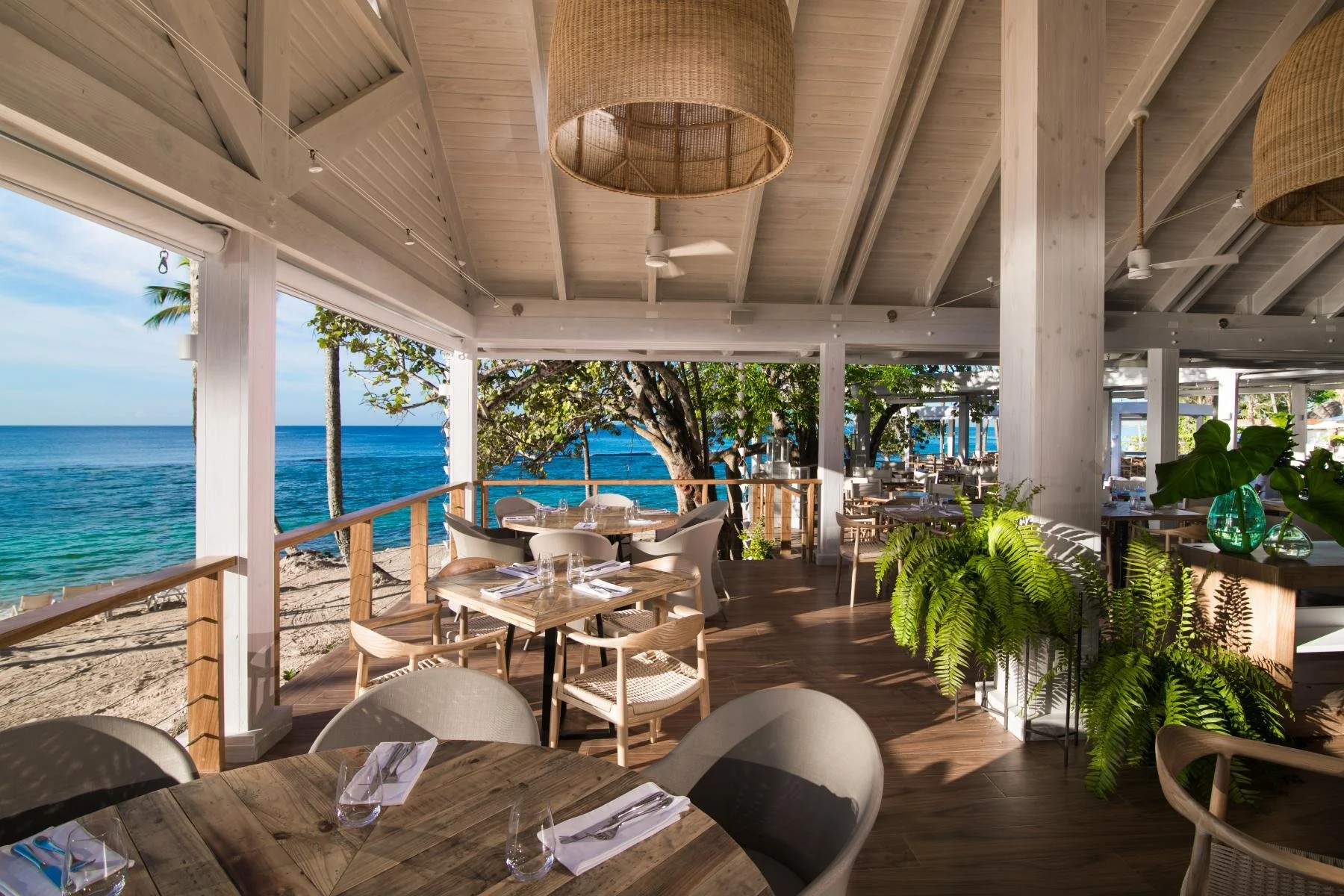 Open-air seaside restaurant with wooden tables and chairs, view of the sandy beach and ocean through open sides, green plants, and trees, with a white ceiling and woven pendant lights.