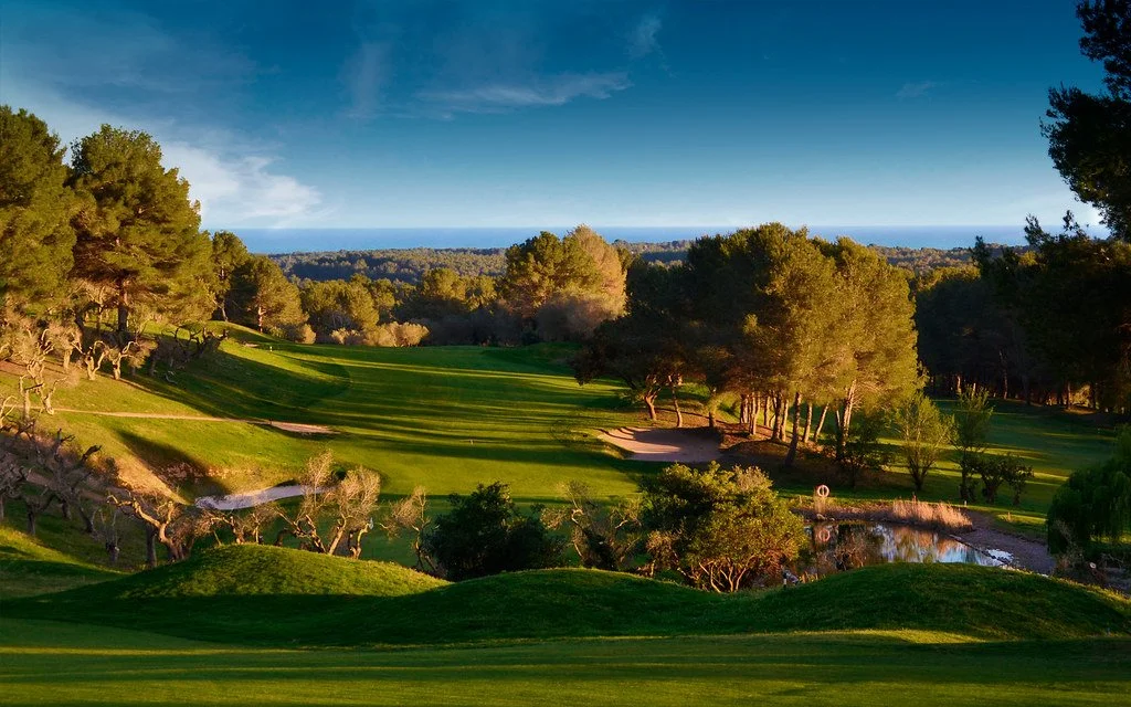 A scenic view of a golf course with lush green grass, trees, water hazards, and sand traps, with the ocean in the distance under a partly cloudy sky.