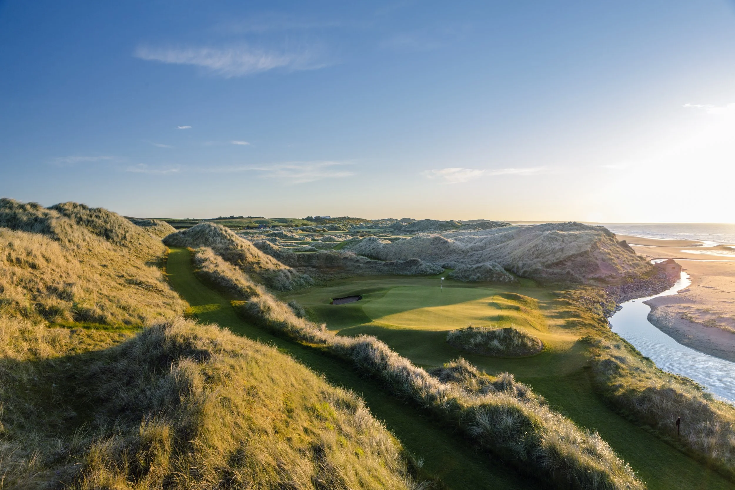 A scenic view of a coastal golf course with lush green fairways and sand dunes, overlooking the ocean at sunset.