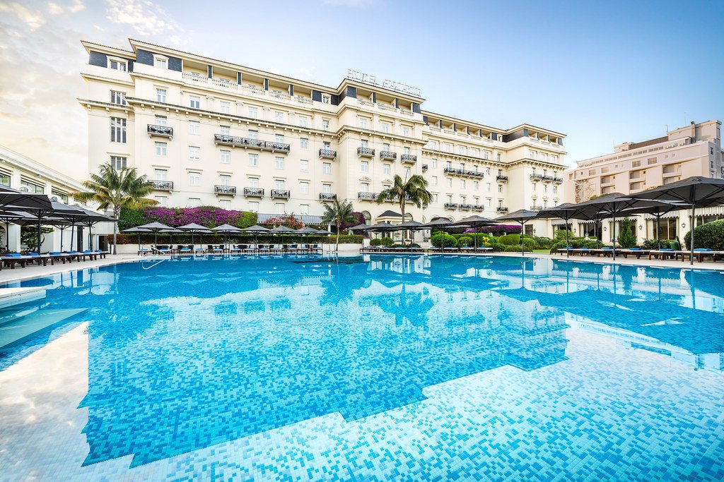 Luxury hotel pool area with blue mosaic pool, surrounding lounge chairs with umbrellas, palm trees, flowers, and a large white hotel building in the background under a clear sky.