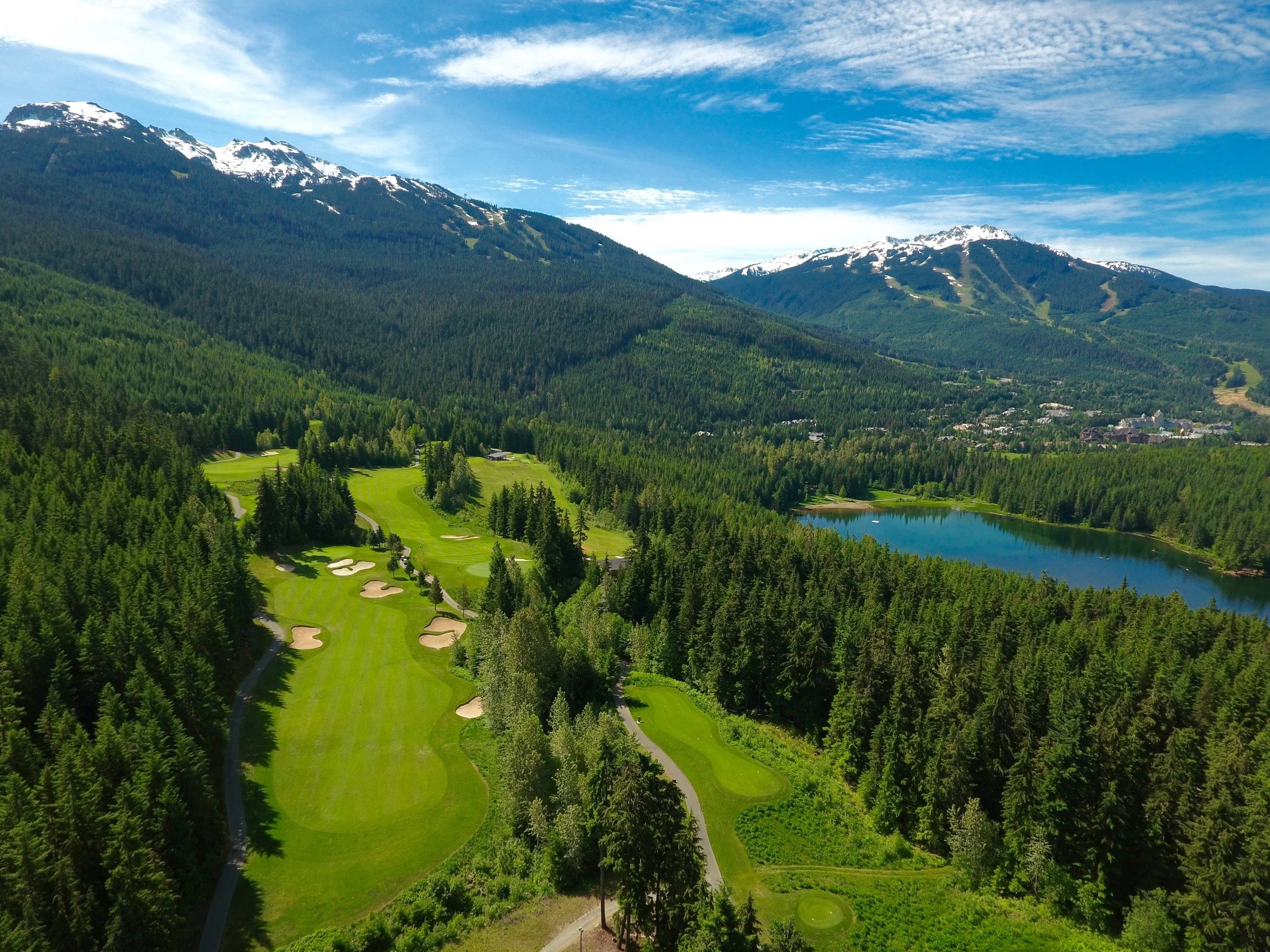 Aerial view of a lush green golf course surrounded by dense forest, with a lake nearby and snow-capped mountains in the background under a blue sky with scattered clouds.