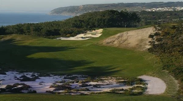 A golf course with green fairways, sand bunkers, and trees, overlooking a body of water and distant cliffs.