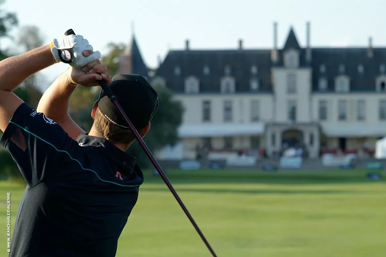 A person in a black cap and black sports shirt preparing to hit a golf ball with a driver on a golf course, with a large hotel or mansion in the background.