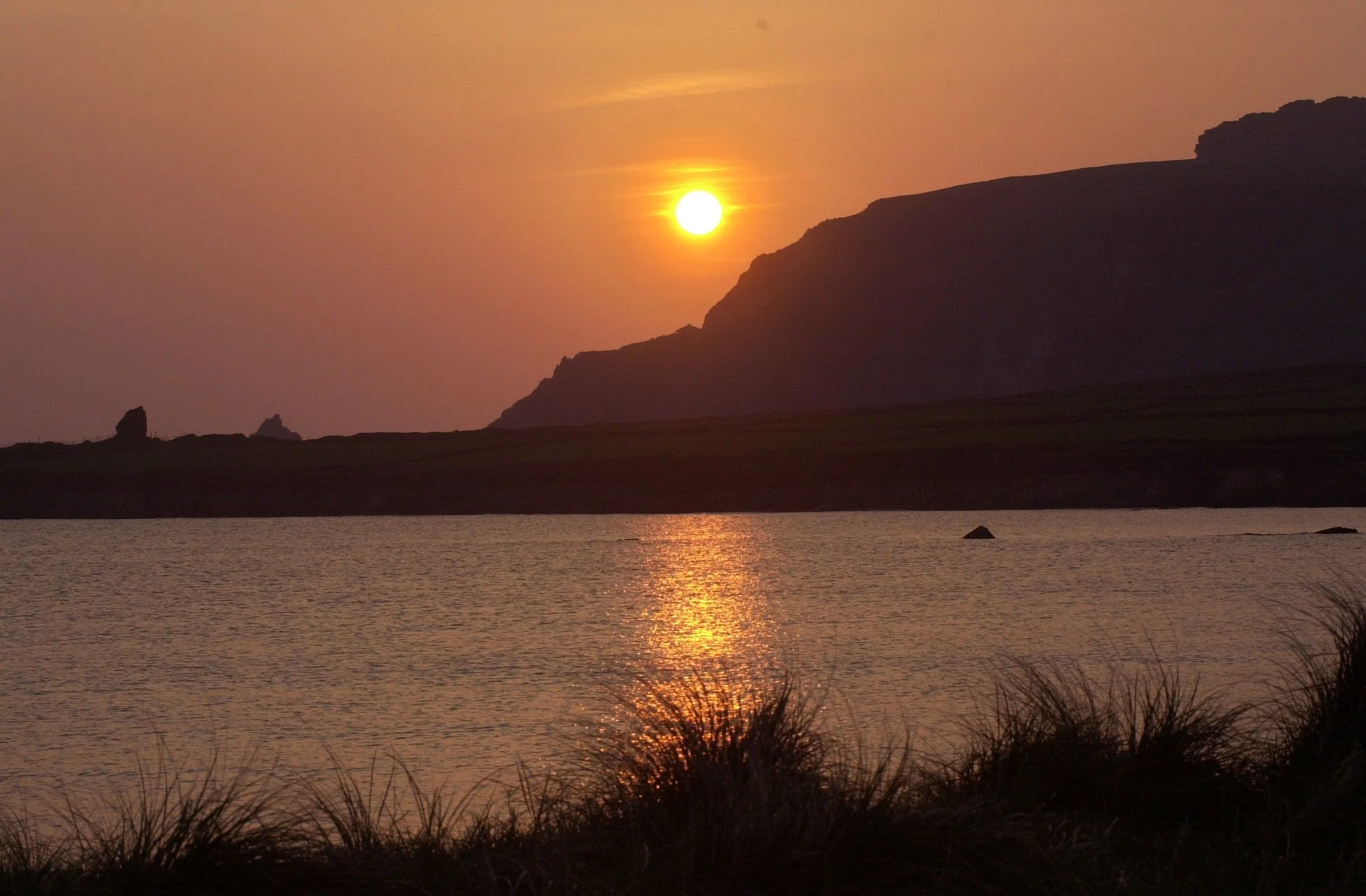 Sunset over a body of water with mountains in the background and grass in the foreground.