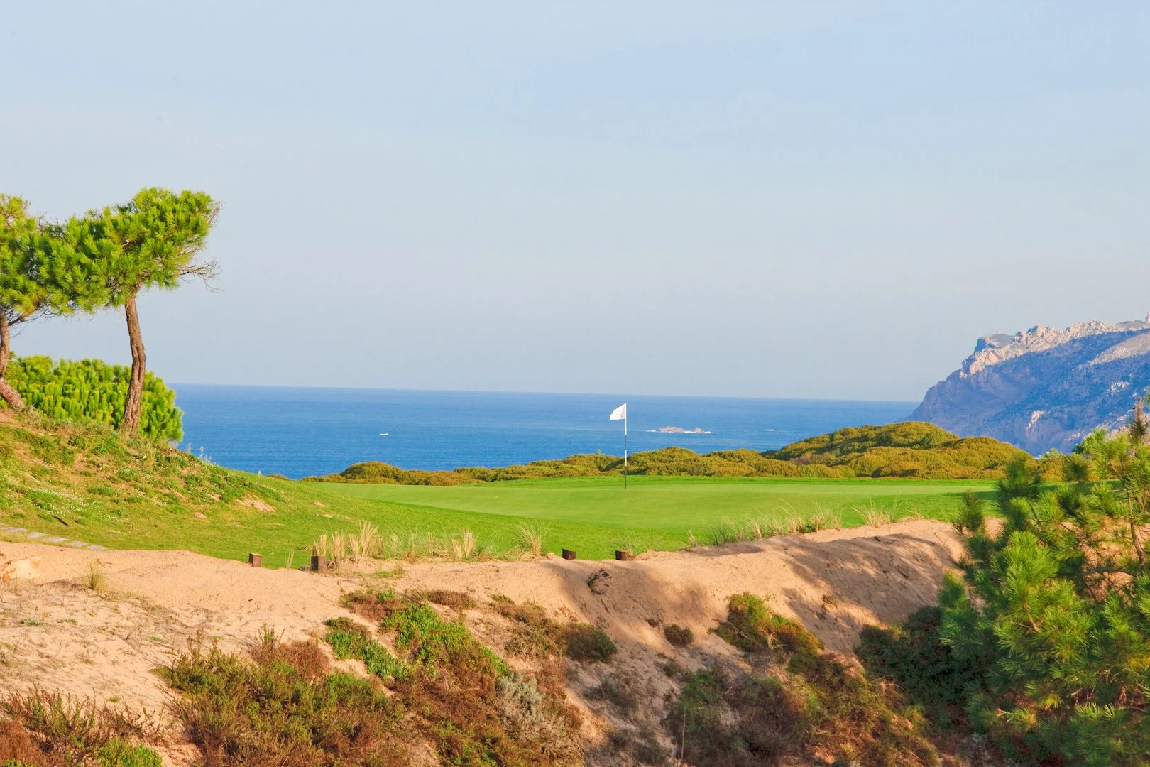 A golf course on the coast with a flag on the green, surrounded by sand and bushes, and overlooking the ocean with mountains in the distance.