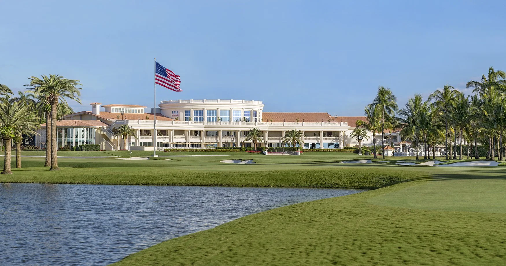 A golf course with a water hazard, green grass, and sand bunkers in the foreground. In the background is a large, white clubhouse with red roof, multiple balconies, and an American flag flying on a flagpole. Tall palm trees line the view.