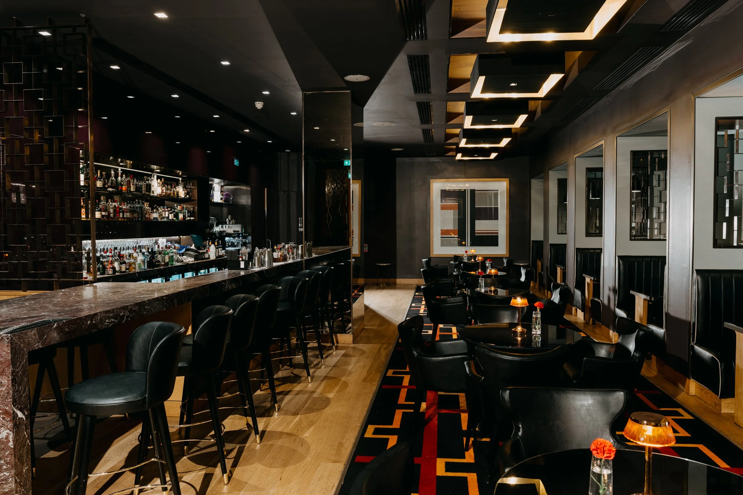 A modern, upscale bar and lounge with black leather chairs, a marble-topped bar counter, and a variety of liquor bottles on display. The space features geometric lighting fixtures on the ceiling, decorative wall art, and dim ambient lighting creating a cozy atmosphere.