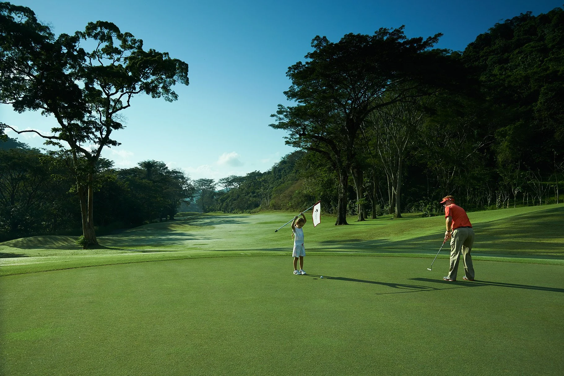 A man and a boy practicing golf on a lush green golf course surrounded by trees, with a clear blue sky overhead.