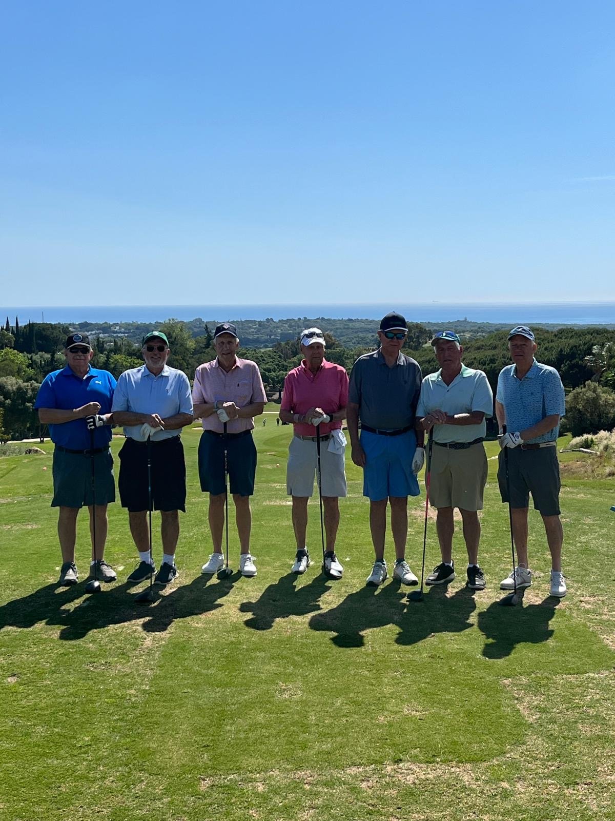 Seven men standing in a row on a golf course, holding golf clubs, with trees, a distant city, and the ocean in the background on a sunny day.