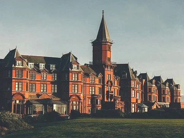 A large, historic red brick building with a tall steeple, featuring multiple turrets and ornate architectural details, set against a clear sky.
