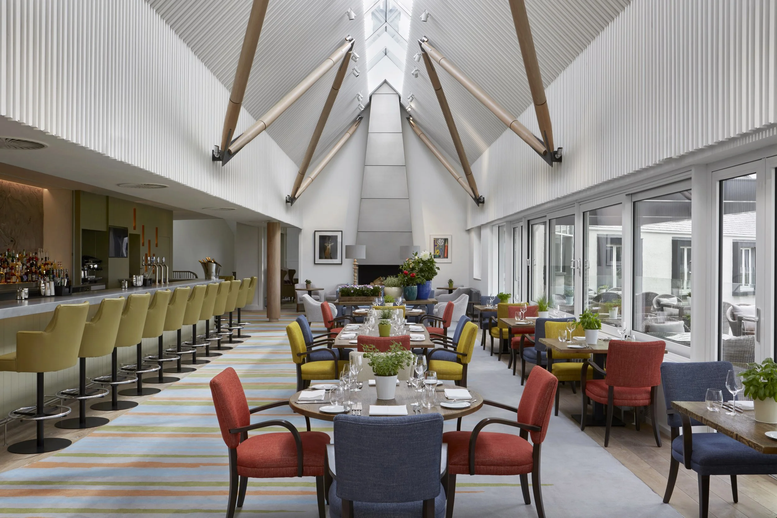 Interior of a modern restaurant with colorful chairs, tables set with glassware, and large windows showing outdoor seating, with an angled ceiling and decorative beams.