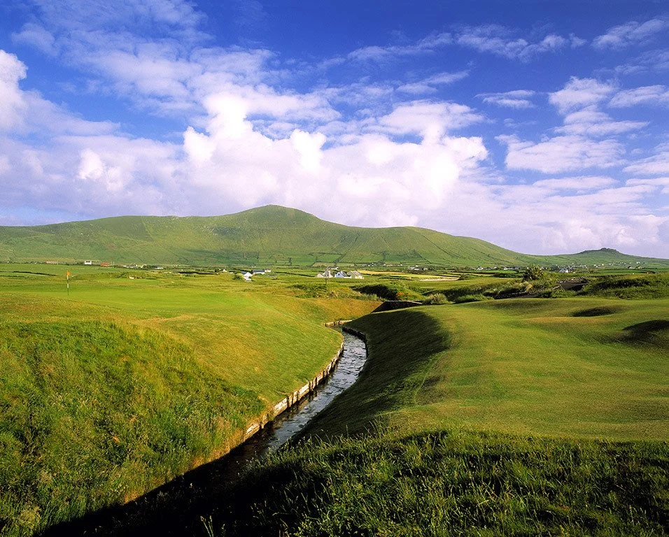 A scenic landscape featuring green rolling hills, a small stream, and a partly cloudy blue sky with scattered clouds.