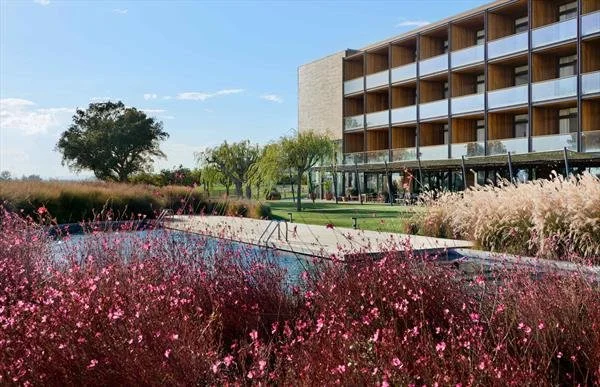 Modern hotel or apartment building with balconies, surrounded by landscaping including pink flowering plants, tall grasses, and trees, near a swimming pool.