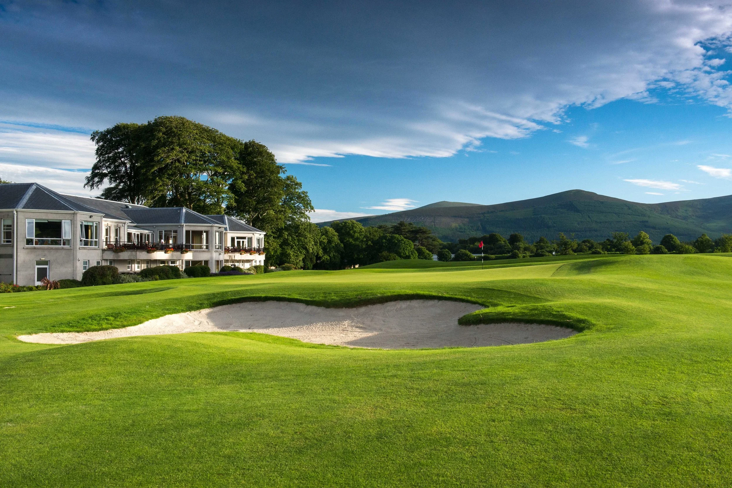 A golf course with a green, sand trap, and flag, surrounded by trees and mountains, with a residential building overlooking the course, under a sky with clouds.