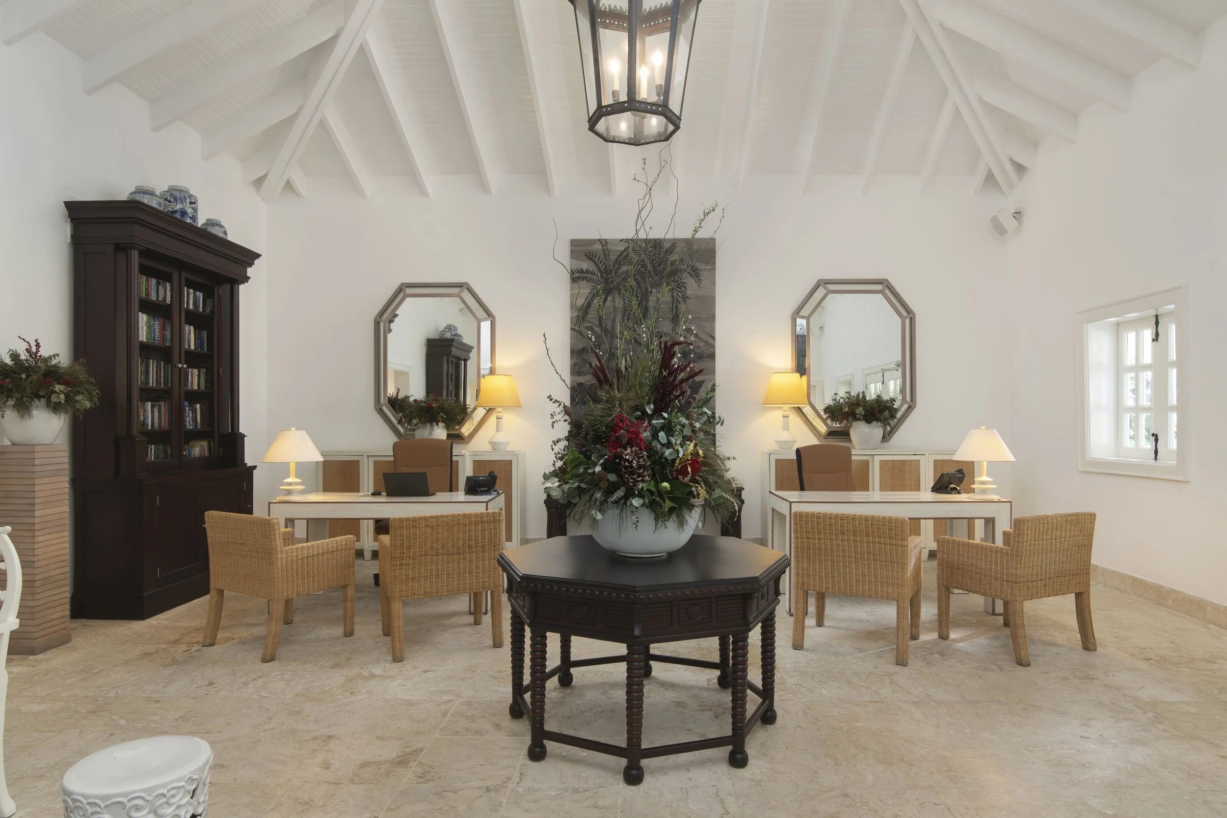 A spacious, well-lit reception area with a high, white vaulted ceiling, a central floral arrangement, two desks with lamps, and a dark wooden bookshelf on the left.