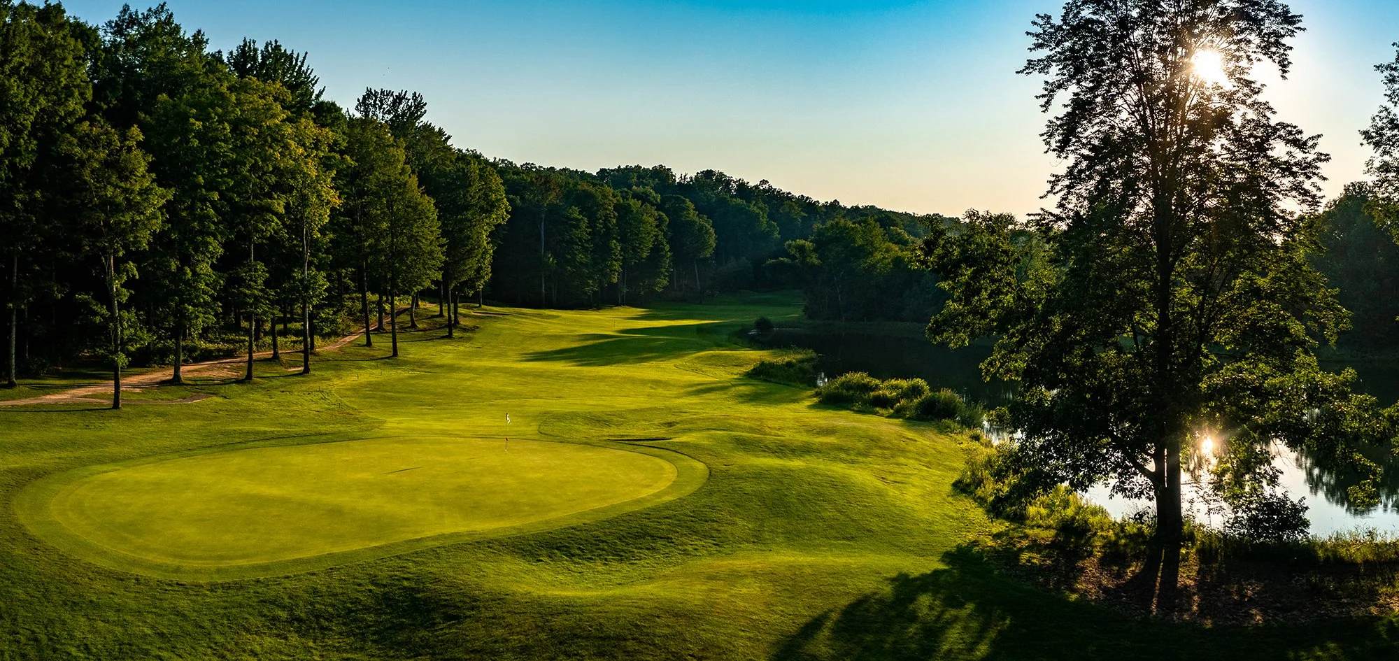 Sunset over a golf course with green grass, trees, and a water hazard.