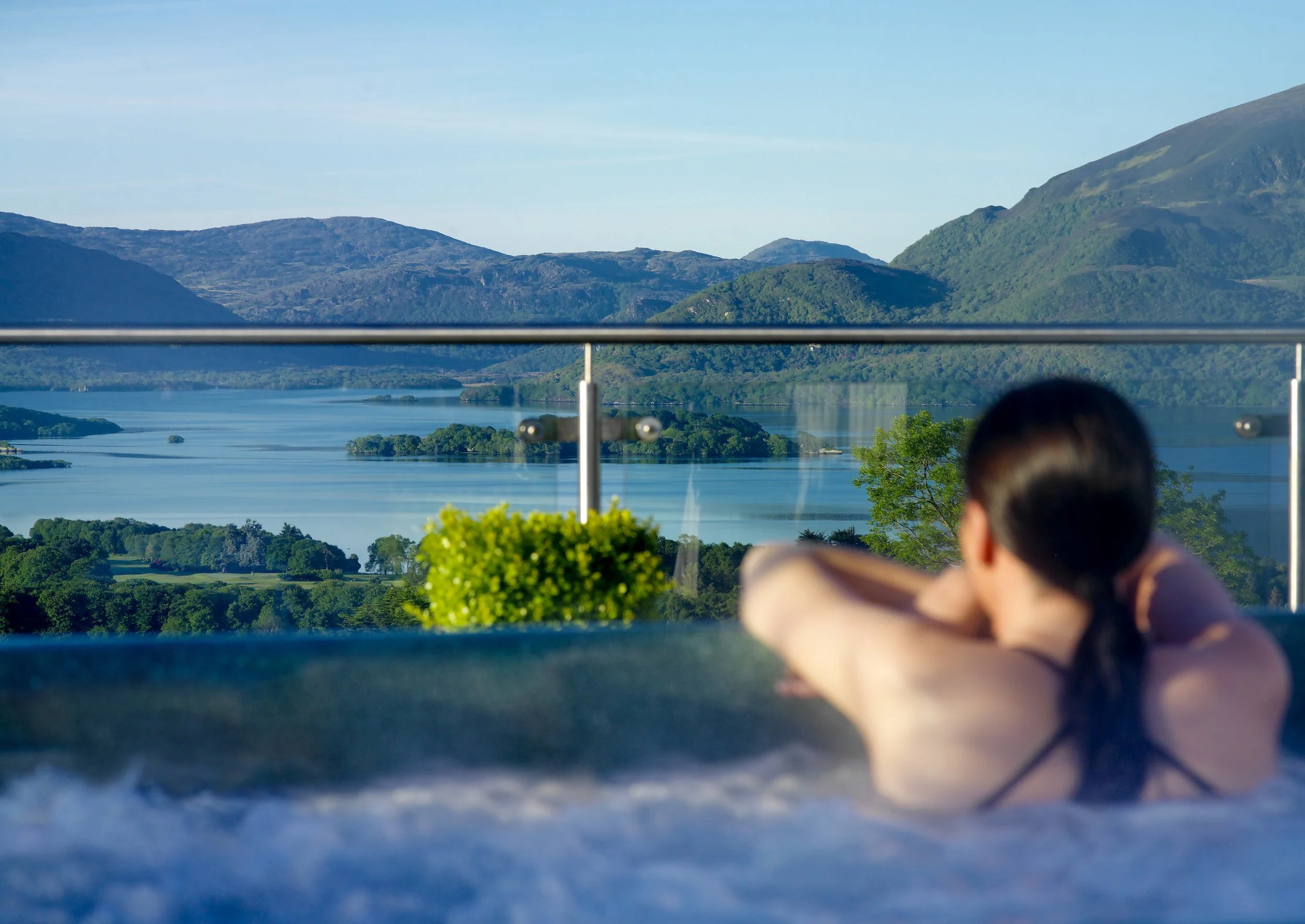 A person with dark hair, wearing a black top, sitting outdoors on a balcony, overlooking a river and mountains during daytime.