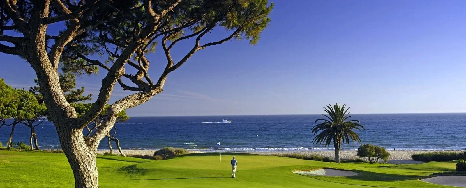 A person standing on a golf course near the ocean with trees, sand traps, and a boat in the water under a clear blue sky.