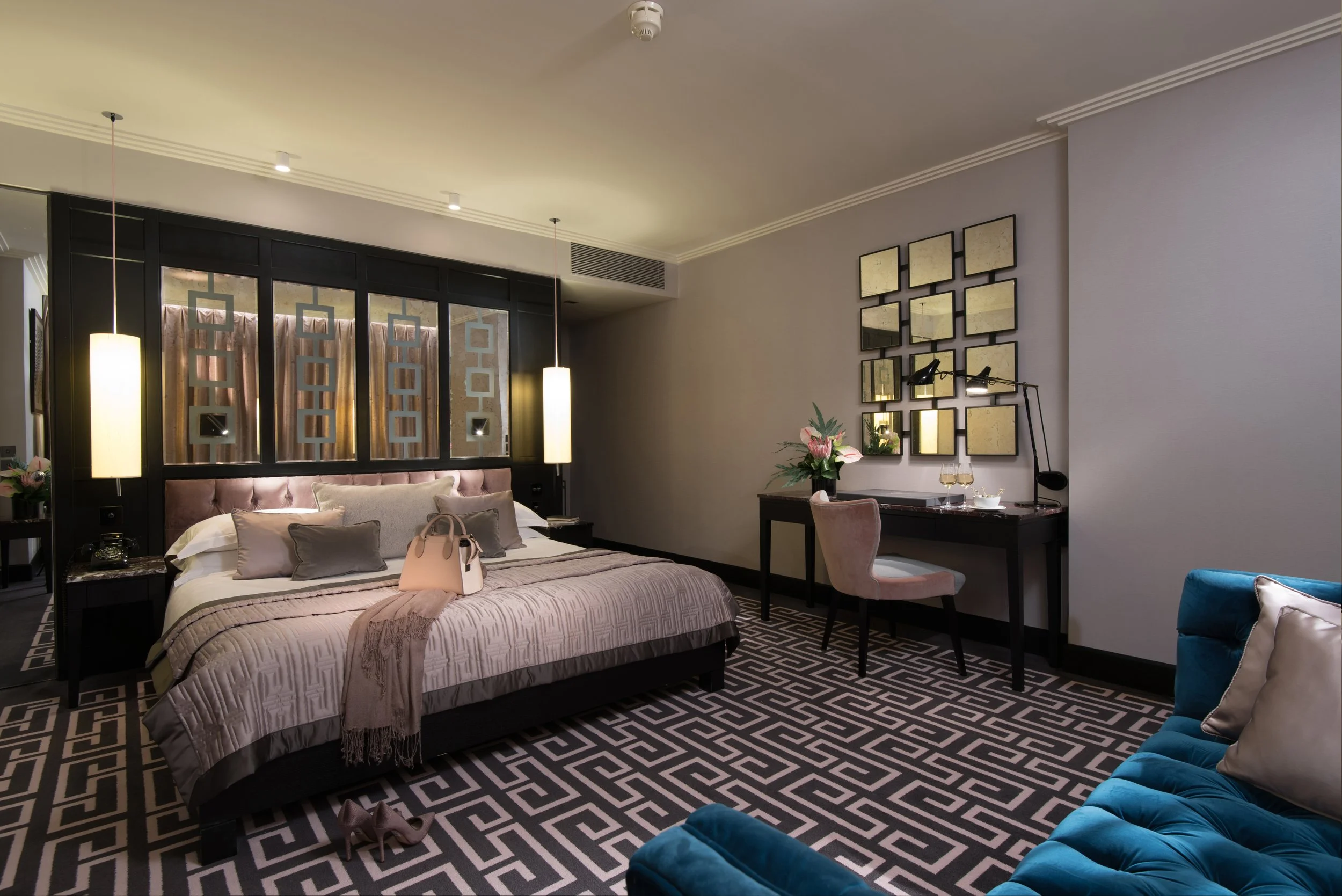 Modern hotel room with a king-sized bed, tufted headboard, multiple pillows, beige bedspread, and slippers. There is a blue velvet sofa, a desk with a chair, a flower arrangement, wall art, and pendant lighting.