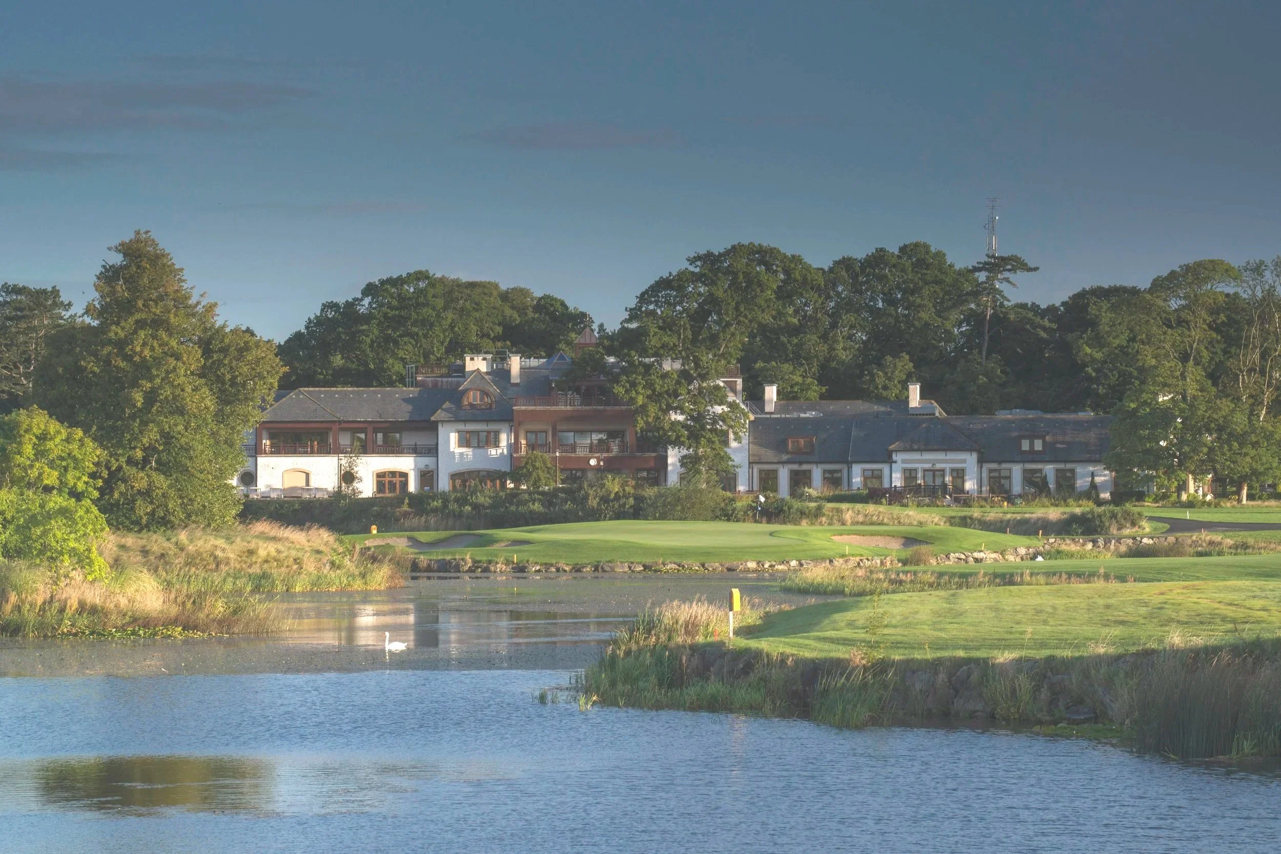A scenic view of a golf course with lush green grass and a water hazard, bordered by trees, with residential buildings in the background under a clear sky.