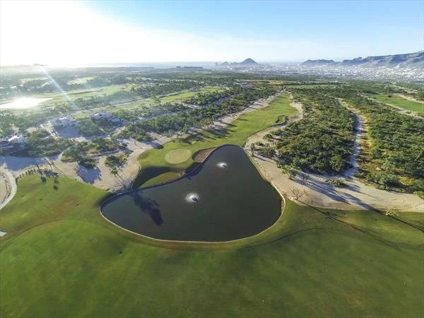 Aerial view of a golf course with a unique water hazard shaped like a heart, surrounded by lush green fairways and trees, with mountains in the distance under a clear blue sky.