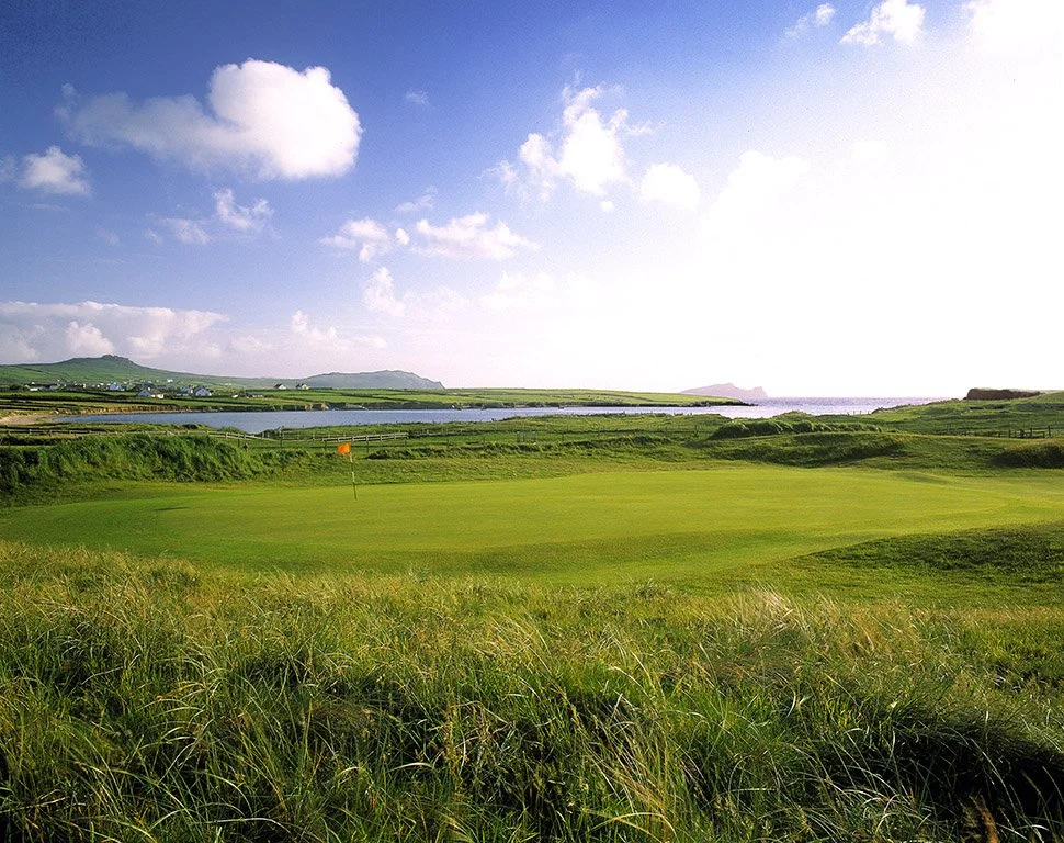 A lush golf course with green fairways, a flag on the green, and water in the background, under a partly cloudy sky.