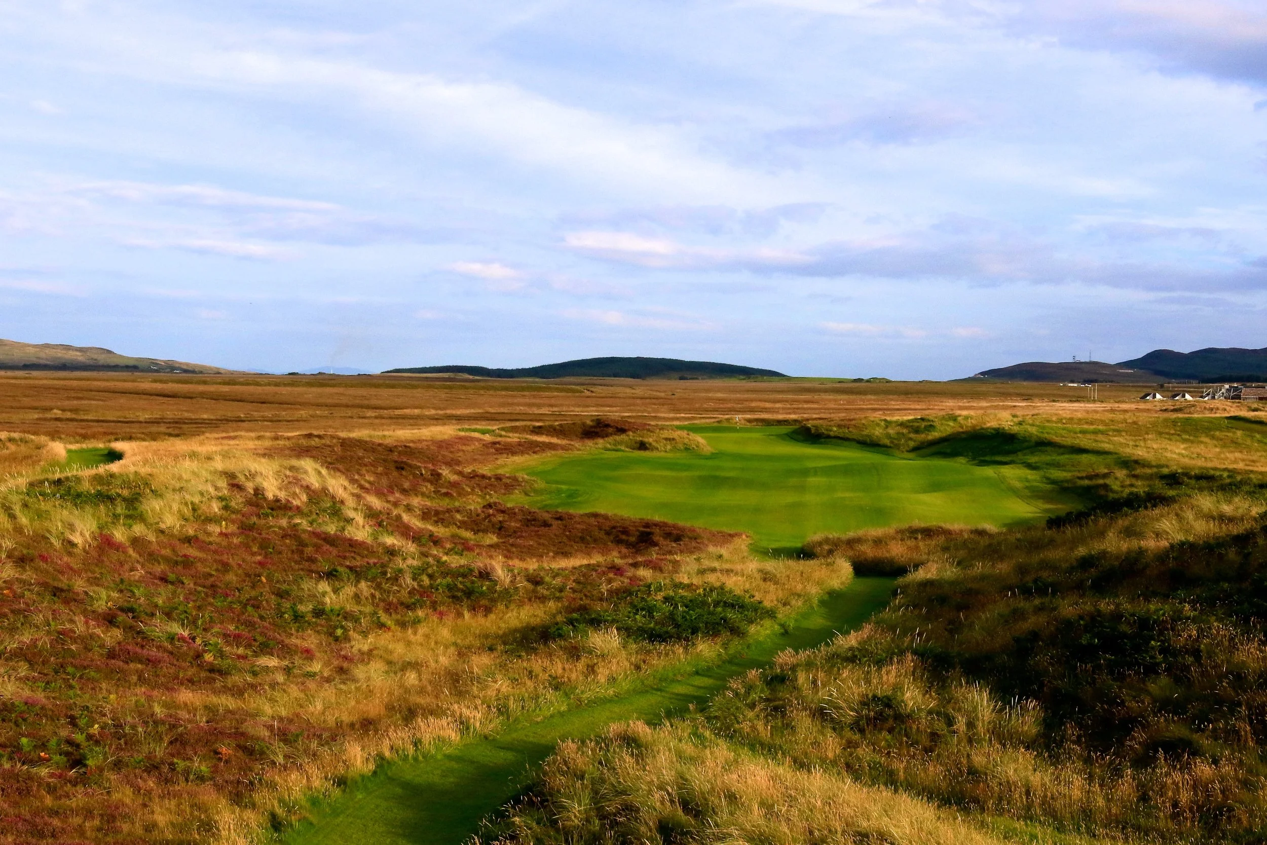A golf course running through a vast open landscape with grassy terrain and hills in the background, under a partly cloudy sky.