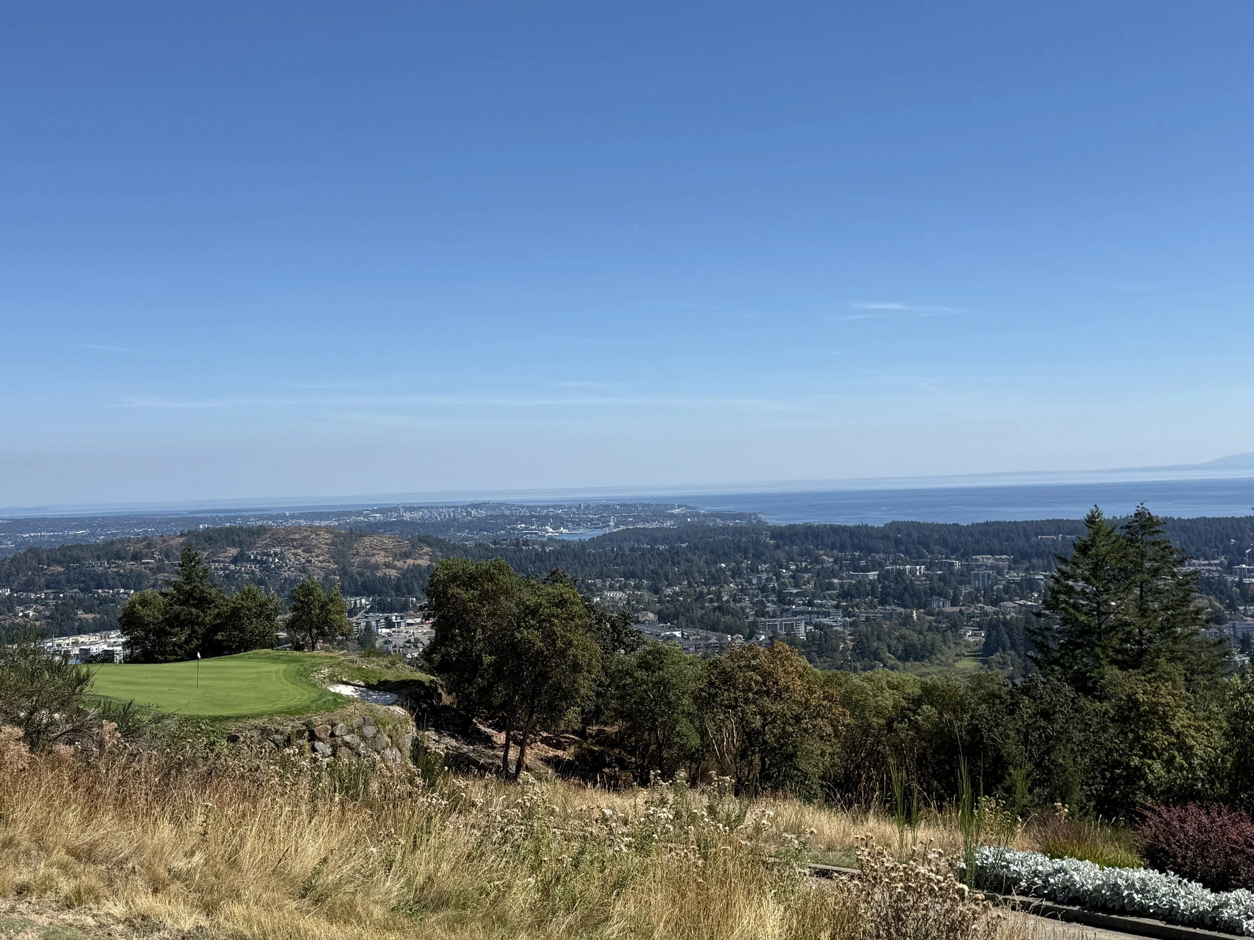 Bear Mountain golf "An elevated panoramic view from a golf resort looking over a vast forested valley towards a coastal city with a deep blue ocean and distant mountains under a clear sky. A manicured green is in the left foreground." }