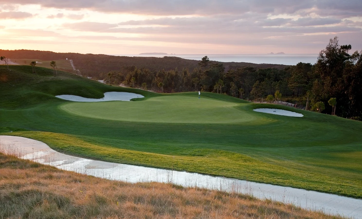 A golf course with green grass, sand traps, and a flag, surrounded by trees and a scenic landscape at sunset.