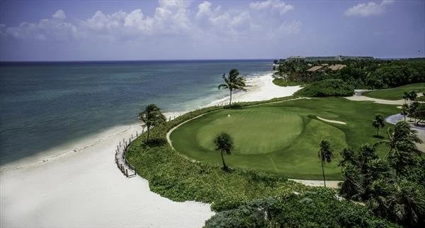 A coastal golf course with green grass, palm trees, and a sandy beach along the shoreline under a partly cloudy sky.