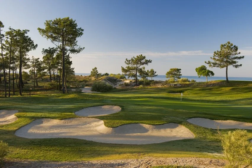 A golf course with sand bunkers, green grass, and trees near a body of water under a blue sky.
