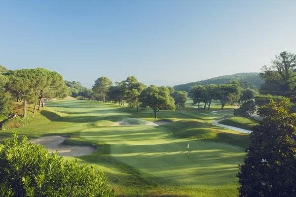 Scenic view of a golf course with lush green fairways, trees, sand bunkers, and a flag marking a hole, under a clear blue sky.