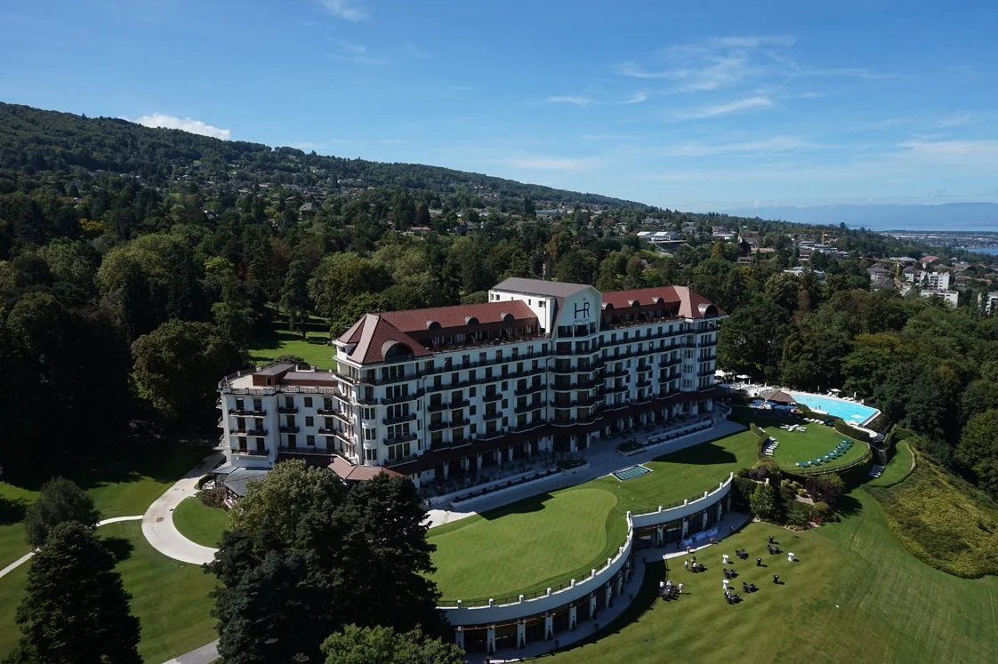 A large multi-story hotel with a red roof, surrounded by greenery and a golf course, with a swimming pool on the right side and a scenic hillside in the background.