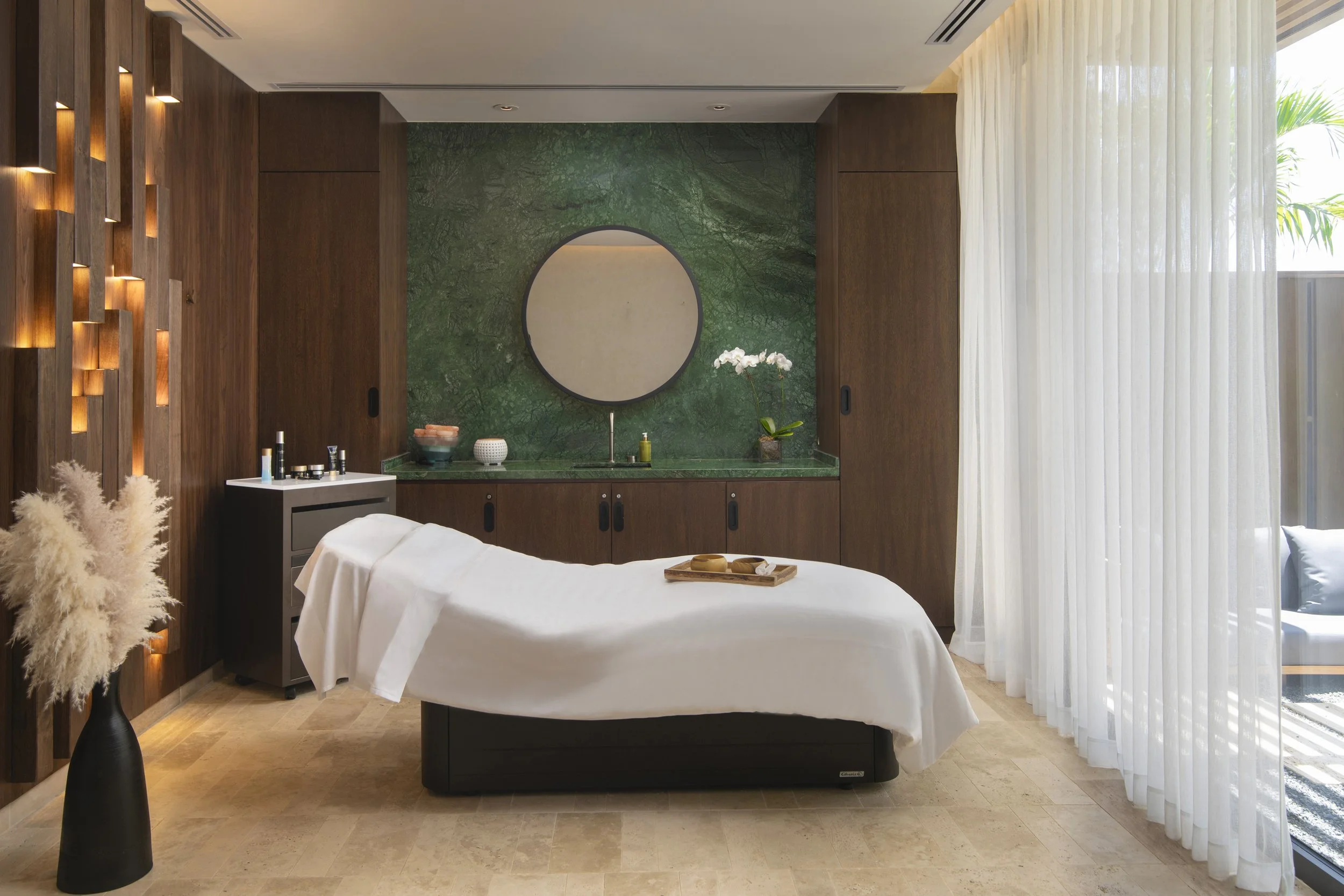 Spa room with massage table, wooden walls, green marble wall with mirror, white curtains, and outdoor seating