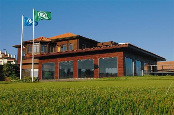 A large building with wooden and brick exterior, floor-to-ceiling windows, and a red tile roof, situated on a well-maintained golf course with green grass and three flagpoles with flags flying.