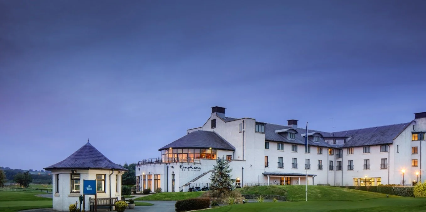 A large white hotel building with multiple floors and windows, situated on a landscaped lawn with a small round building in front. The sky is transitioning from day to night, showing a dusky blue hue.