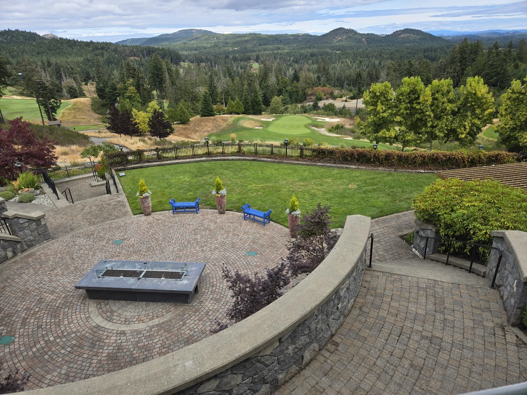 "An elevated view of a circular stone patio with a fire pit, leading to a manicured lawn with bright blue benches. Beyond the lawn, a lush green golf course is nestled among dense forests and rolling hills under a partly cloudy sky."